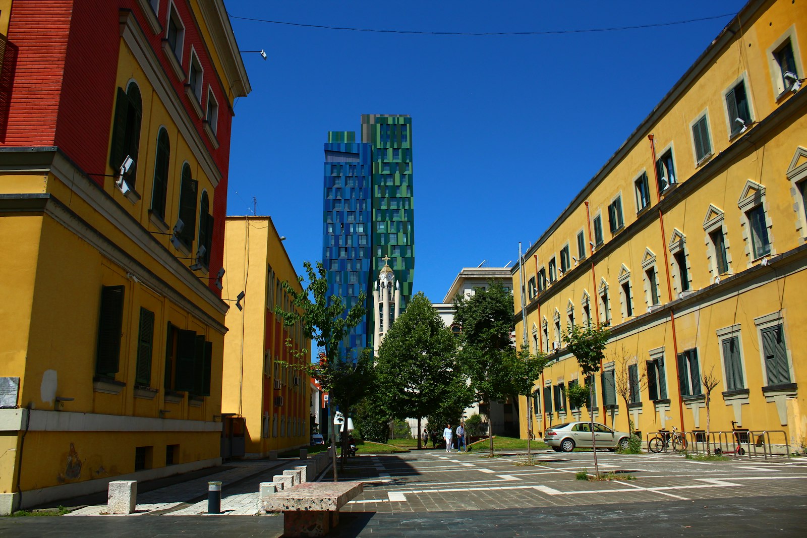 A Tirana street of ochre and red painted facades under a deep blue sky, with the blue-and-green TID Tower rising behind
