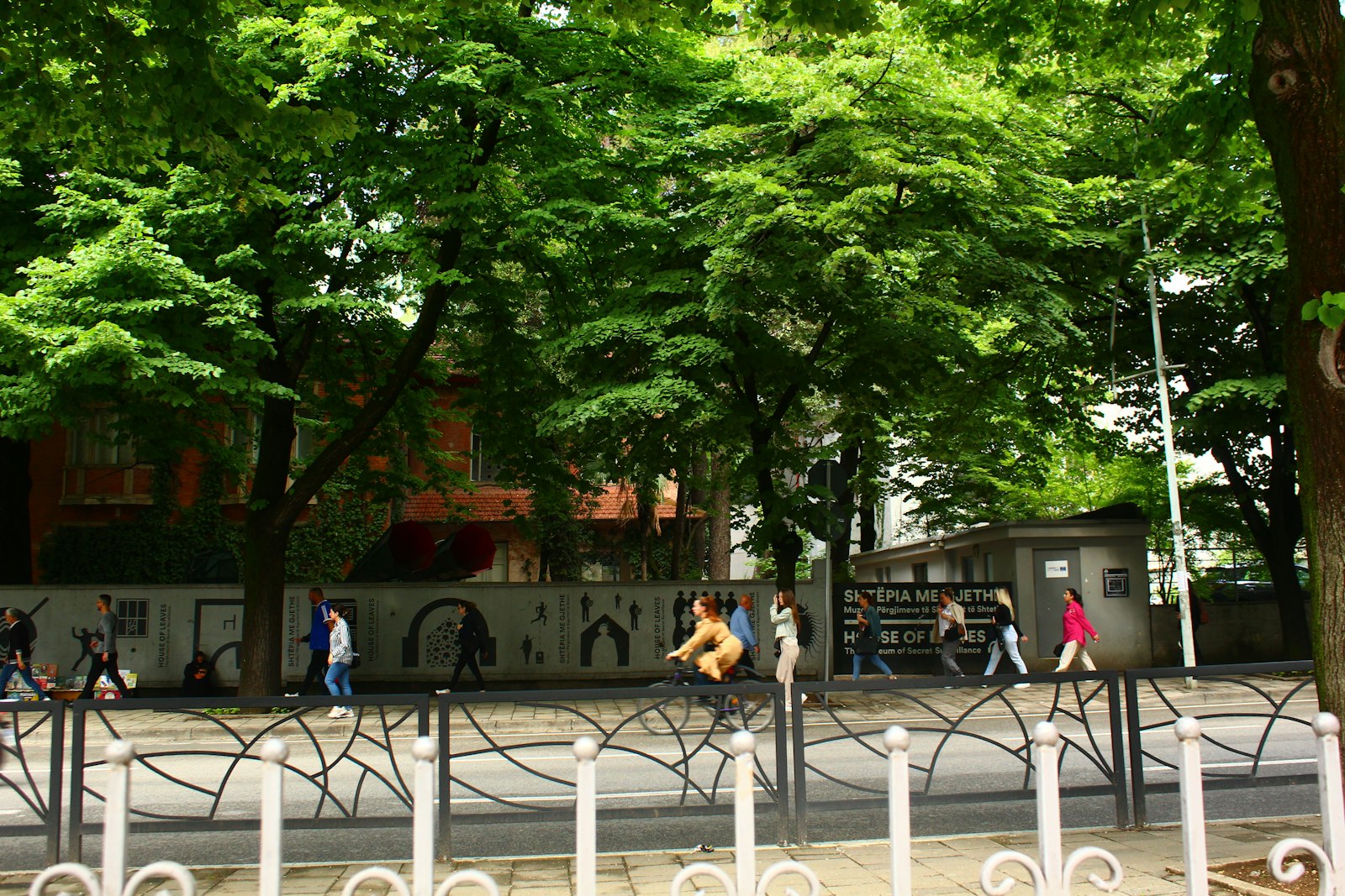 People walking past the House of Leaves museum in Tirana under green street trees